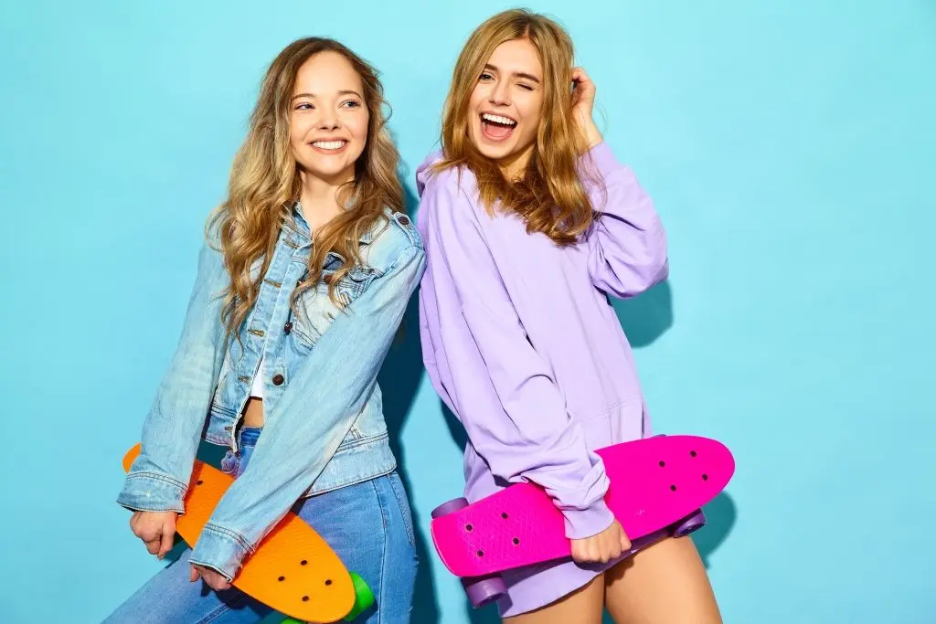 two-young-stylish-smiling-blond-women-with-penny-skateboards-women-in-summer-hipster-sport-clothes-posing-near-blue-wall-positive-models.jpg